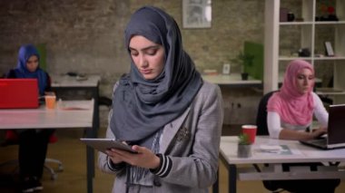 Şirin Arapça bir kadın gri hijab içinde swwiping onu gerey tablet ve diğer İslam çalışan kadınların yanında dururken presicely bakarak.