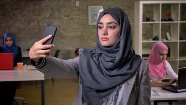 Perempuan lucu arab mengenakan jilbab abu-abu berdiri dan memegang ponselnya sementara gadis-gadis lain yang mengenakan jilbab menggunakan komputer mereka di kantor bata modern — Stok Video