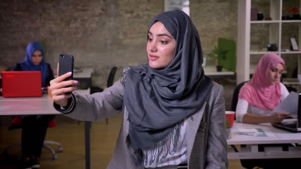 Amazing arab female is talking over phone, having wvideo call while standing in brick studio with working middle east girls sitting behind her — Stock Video