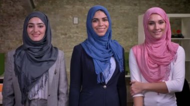 Üç Arap kadın hijab giyen gülümseyen, pembe, mavi ve gri ayakta ve modern tuğla ofis, fotoğraf bakarak