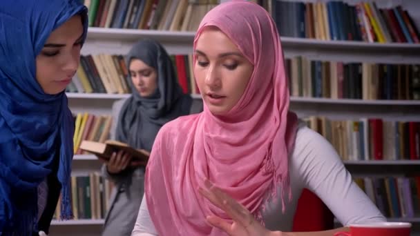 Belle donne arabe stanno parlando e seduti a tavola insieme, avendo conversazione, indossando hijab, libro di lettura ragazza del Medio Oriente sullo sfondo — Video Stock