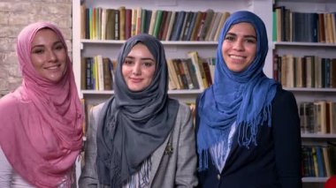 Üç ayakta Türban Arap kadın büyük bir güven ile başlayan ve gülümseyen, Kütüphane arka plan, çok renkli kitap kameraya arıyoruz