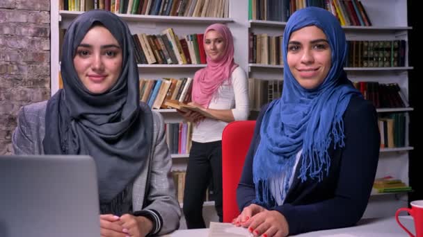Απολαυστική πρόσωπα του hijab μουσουλμανική κορίτσια που κάθονταν σε βιβλιοθήκη κοντά το laptop και κοιτάζοντας με χαμογελαστά πρόσωπα φωτογραφική μηχανή, εικόνα της σύγχρονης ζωής — Αρχείο Βίντεο