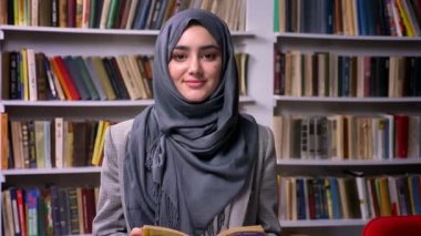 Güzel gri hijab zevkli oldukça Arap kız kitap raf önünde duruyor ve kamera chill gülümseyen, rahat vibes