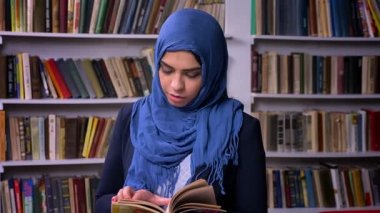 Koyu mavi hijab içinde harika Arap kadın kitap kütüphane, sakince ayakta ve kendinden emin kamera, sakin vibes bakışta tutuyor