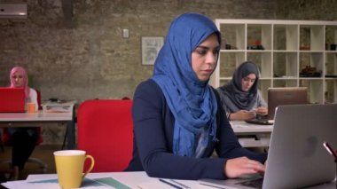Koyu mavi hijab Akıllı güzel çalışan arap kız masaüstünde oturuyor ve onun bilgisayarda yazarak, kapı, tuğla arka plan meslektaşları ile çevrili