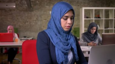 Muhteşem müslüman kadın oturuyor ve arka planda çalışan Arap kadınlar ile dizüstü bilgisayarda yazarak, kamera düz bakmak, sakin yüz