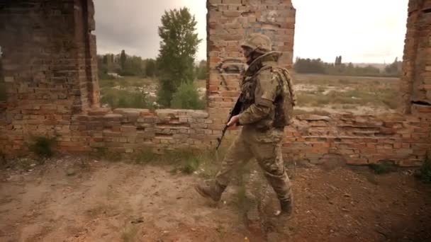 Pejuang berjalan dengan kamuflase menyeberangi bangunan bata yang ditinggalkan di luar dengan senjata otomatis di lengannya, percaya diri dan orang yang kuat — Stok Video