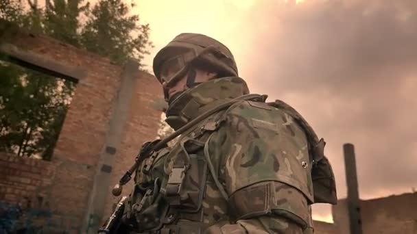 Petugas kaukasia yang gigih berjalan dengan tenang melalui bangunan bata yang ditinggalkan dan memegang senjata otomatis, langit mendung gelap, pembela ilustrasi — Stok Video
