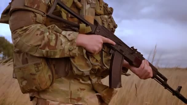 L'homme militaire tient l'arme et fait des pas confiants à travers l'herbe verte supérieure à l'extérieur, un ciel sombre, défendant une position authentique — Video