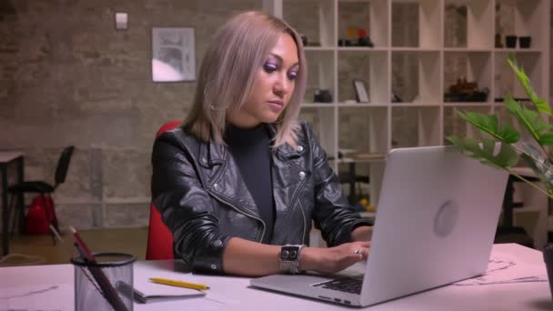 Linda mujer rubia caucásica reflexiva está escribiendo en el teclado mientras está sentada aislada en su lugar de trabajo en interiores — Vídeos de Stock