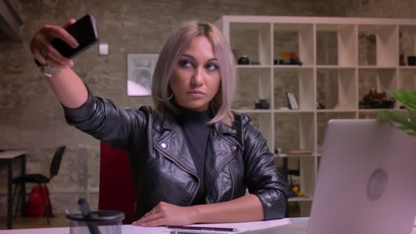 Słodkie entuzjastyczne kaukaski blond kobieta w robienia selfie na jej telefon, siedząc przy pulpicie siedząc kryty chill, nowoczesne ilustracja, tło office — Wideo stockowe