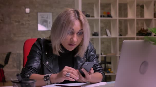 Fantastisk blondin kaukasiska kvinna tittar just på sin telefon och röra skärmen sittande på arbetsplatser inomhus, isolerade i ljus office tegel bakgrund, moderna verkligheten — Stockvideo