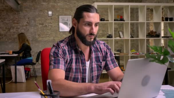 Mignon homme d'affaires caucasien concentré est en tenue décontractée et avec la barbe noire est dactylographier sur ordinateur portable tout en étant assis dans un bureau en brique légère, illustration moderne — Video