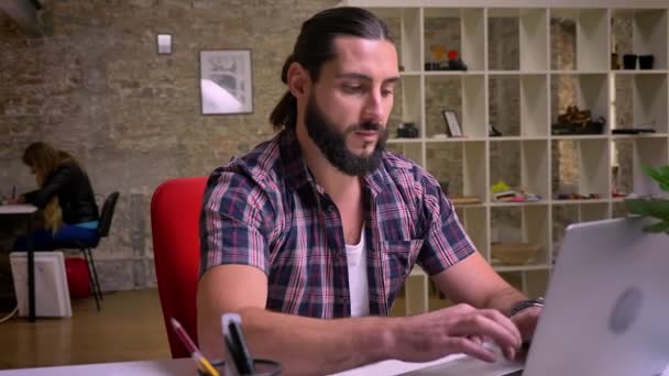 Joli mec caucasien avec une grande barbe noire tapant sur le clavier tout en étant à son bureau, puis en regardant avec un sourire à la caméra, bureau moderne de nos jours — Video