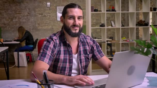 Sonriente hombre caucásico con barba bonita y pelo largo es relajante durante el tiempo de trabajo y mirando a la cámara con sonrisa mientras está sentado en su lugar de trabajo en la oficina moderna aislado — Vídeo de stock