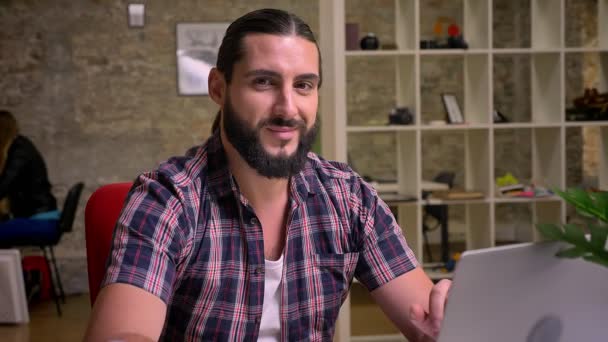 Felicidad sin la sonrisa del hombre guapo caucásico con barba negra que está sentado cerca de la computadora portátil en el escritorio en la oficina de ladrillo y se concentró en su trabajo, ilustración moderna interior — Vídeo de stock