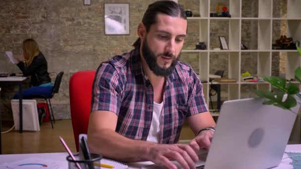 Disfrutable hombre caucásico con barba negra se ríe mientras mira su computadora y escribe, se divierte en el lugar de trabajo, estilo casual un hombre relajado, estudio de ladrillo insode — Vídeos de Stock