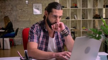 Siyah sakallı ve uzun saç beyaz erkek çalışan, yakışıklı klavyede yazarak o zaman düşünceli yüzüne tuğla ışık arka plan üzerinde izole onun okul sýrasý ile oturuyor ve