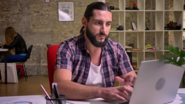 Memnun yakışıklı beyaz erkek siyah uzun sakallı ve laptop yazarak ve gururla bakarak saç tuğla modern ofiste otururken yüzüdür