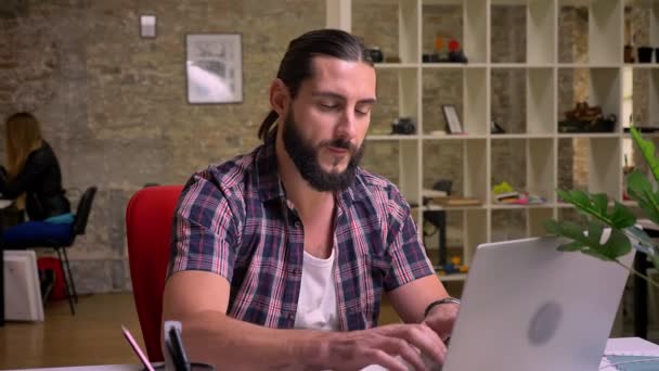 Digitar bom homem caucasiano com grande barba está sorrindo confiantemente enquanto digita em seu computador na área de trabalho na sala de luz de tijolo, hoje em dia ilustração de trabalho — Vídeo de Stock