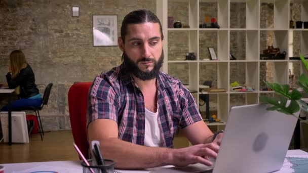 Regard douteux réfléchi de l'homme caucasien à barbe noire qui regarde bizarrement l'écran de son ordinateur portable et assis à son lieu de travail isolé sur fond de brique — Video
