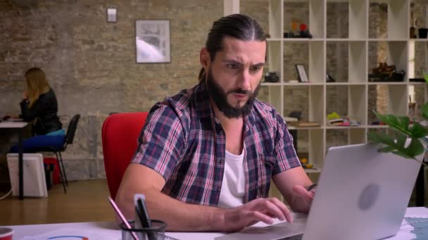 Beautiful fit man, caucasian appearence with black beard is sitting at workplace and typing on computer with small pauses laughing and looking at ther screen, cherfully, modern light office on — Stock Video