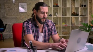 Güzel beyaz sakallı adam hayal kırıklığı kollarıyla süre işyerinde oturan ve Laptop kapalı, diğer meslektaşları arasında yazarak gösteriyor