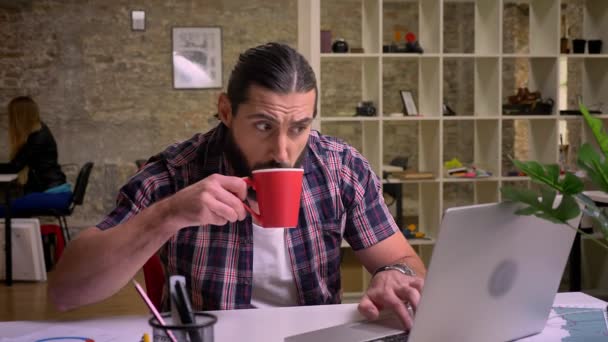 Hombre caucásico concentrado con el pelo negro largo y la barba está escribiendo en el ordenador portátil y beber de la taza roja mientras que cuelga en su lugar de trabajo, ilustración moderna de la oficina — Vídeo de stock