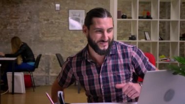 Güzel siyah sakallı mutlu genç bir yaratıcı işçi vasıl okul sırası oturan ve görüntülü görüşme sahip dizüstü bilgisayar, tuğla modern ofis