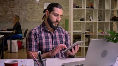 Sakin yaratıcı casual beyaz erkek uzun saç ve siyah sakallı ile onun tablet kapalı, Masaüstü, dizüstü bilgisayar, tuğla ofis arka plan arkasında otururken swiping