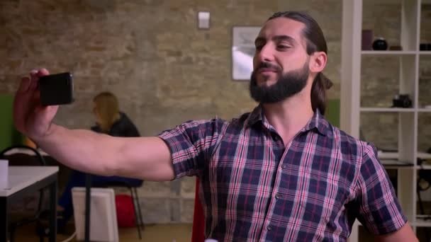 Felice ragazzo caucasico carino con la barba nera sta parlando selfie sul suo telefono mentre seduto sul desktop vicino alle attrezzature di lavoro, muro di mattoni sullo sfondo — Video Stock