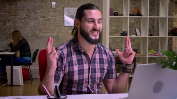 Kaukaski szczęśliwy entuzjastycznie modelu z czarną brodą jest wykazanie z gestem, siedzi jego kciuki, Cegła nowoczesne biuro tło i laptop za nim — Wideo stockowe