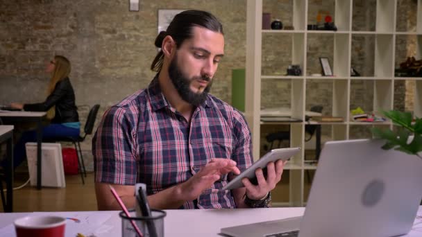 Calma creativa caucásico casual masculino con el pelo largo y la barba negra está deslizando su tableta mientras está sentado en el interior en el escritorio, detrás de la computadora portátil, fondo de la oficina de ladrillo — Vídeo de stock