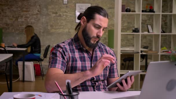 Απολαυστική γενειοφόρος άνδρας Καυκάσιος χρησιμοποιεί tablet ήρεμα και εστιασμένη ενώ κάθεται στο χώρο εργασίας, υπερήφανο πρόσωπο και ατμόσφαιρα ψύξης, εσωτερική απεικόνιση — Αρχείο Βίντεο
