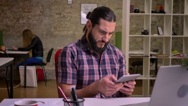 Visage énergique de caucasien heureux, mâle avec une belle barbe noire, regardant l'écran de la tablette, le glissant tout en étant assis dans un bureau moderne en brique — Video