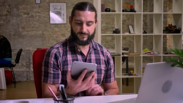 Bonita sonrisa feliz del hombre caucásico con barba que está mostrando la pantalla verde en su tableta y mirando a la cámara recta mientras está sentado en el escritorio, persona de trabajo en el fondo, estudio de ladrillo — Vídeo de stock