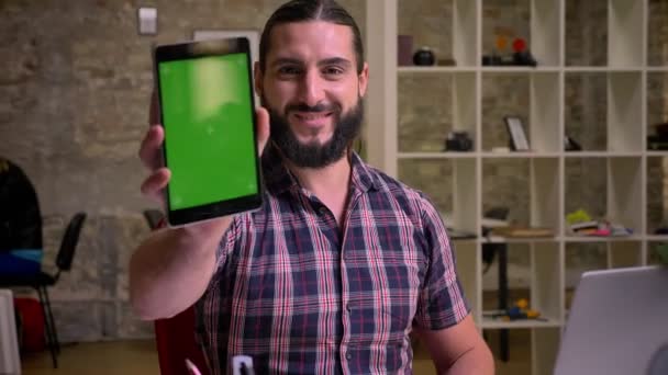 Trevligt leende ansikte kaukasiska affärsman med svart skägg som är visar tablett med grön skärm medan du sitter på sin arbetsplats i moderna ljusa kontor isolerade — Stockvideo