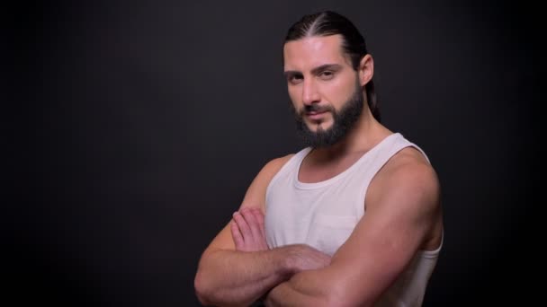 Muscular caucasian bodybuilder with black nice beard is looking seriously at camera and stadnign on black background isolated and calm, whle wearing and strond arms — Stock Video