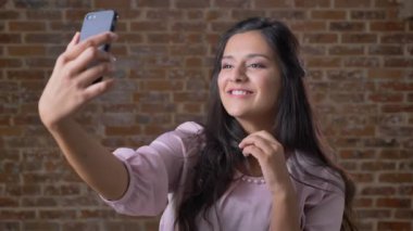 Talike selfies iki parmak gösterilen ve kahverengi Studio, tuğla arka plan dururken gülümseyerek telefonu ile mutlu siyah saçlı beyaz kız ürpertici olduğunu