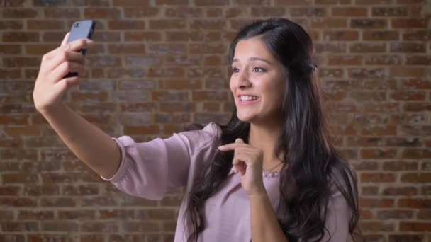 Χαρούμενη μιλάμε καυκάσιος γυναίκα κρατά smartphone στο ένα χέρι και έχοντας κλήση βίντεο, στέκεται χαλαρή, τούβλο πίσω, καφέ στούντιο — Αρχείο Βίντεο