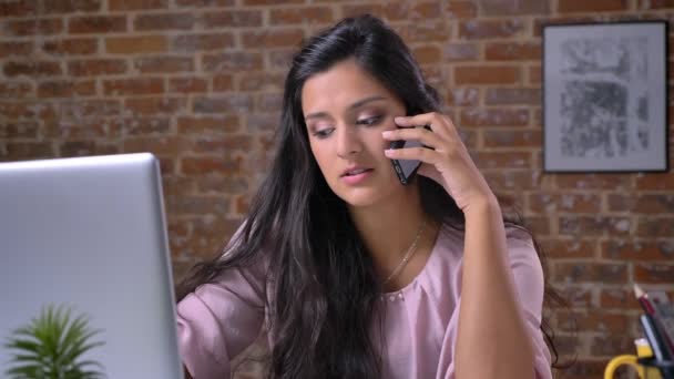 Bonita mulher de negócios caucasiana está falando por telefone com confiança enquanto sentado e olhando para tela og laptop, estar em estúdio de tijolo, trabalhando ilustração — Vídeo de Stock