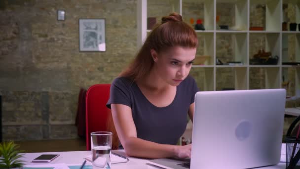 Nice red haired girl is sitting and working on computer and then looking happily at camera, cheerful smile and natural cute face, modern office — Stock Video