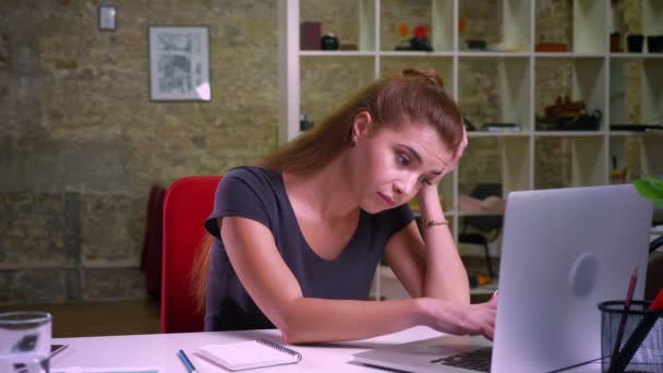 Kızıl saçlı akıllı güzel çalışma beyaz kadın otururken dizüstü bilgisayar ve ofis donanımları, masaüstü kapalı, uzun saç ve güzel görünüm yakınındaki esniyor — Stok video