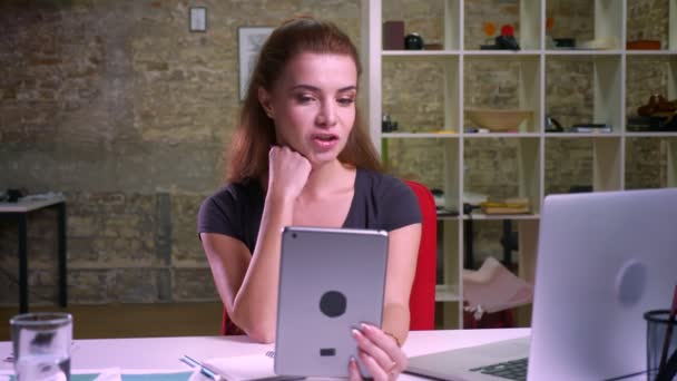 Симпатичная имбирная кавказка получает видеозвонок за планшет, сидя за компьютером, расслабляясь, участвуя в своем рабочем процессе — стоковое видео