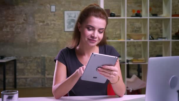 Mulher branca bonito está mostrando tela azul no tablet e sorrindo alegremente para a câmera enquanto sentado no escritório de tijolo brilhante na área de trabalho — Vídeo de Stock