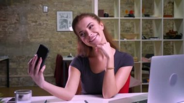 Mutlu yüz ve keyifli telefonla fotoğraf çekmek ve iki parmak neşeyle modern ofiste otururken gösterilen sevimli beyaz Kızıl saçlı kız jesti