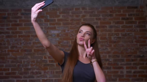 Cute caucasian female is taking selfie and showing her two fingers on her phone while standing on brick background, happy face and smiling person — Stock Video