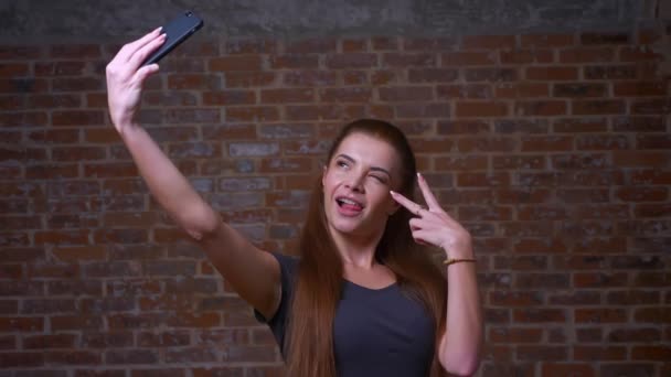 Erstaunliche kaukasische Ingwerweibchen zeigt ihr Glück, während sie Selfie-Fotos mit der Webkamera ihres Smartphones macht, während sie steht, entspannt ist, im Backsteinstudio chillt — Stockvideo