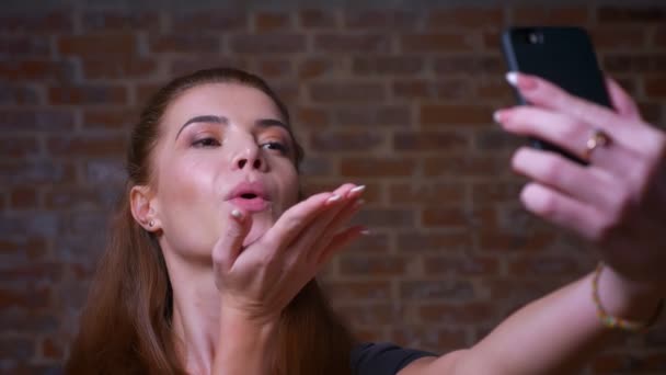 Piuttosto caucasico zenzero donna primo piano è l'invio di baci e soffia a macchina fotografica, parlando selfie guardando il suo telefono concentrato, sorriso felice su sfondo mattone — Video Stock